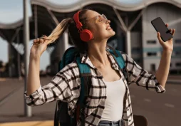 Pessoa jovem com mochila e fones de ouvido vermelhos segurando smartphone enquanto escuta música ao ar livre em frente a aeroporto, conceito de viagem e programa de milhas.