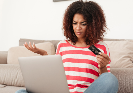Mulher com expressão de dúvida segurando cartão de crédito enquanto usa notebook, representando problema com pagamento ou transação recusada online.
